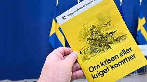 mano sosteniendo la guía oficial "Si llega la crisis o la guerra" sobre un fondo de banderas suecas