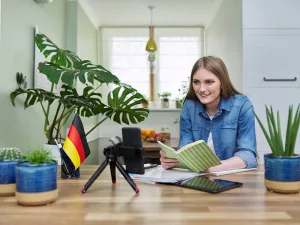 Remote distance learning, online lesson of female student sitting at home using smartphone on tripod. On table textbooks, notebooks, flag of Germany. Female teaching, learning German language